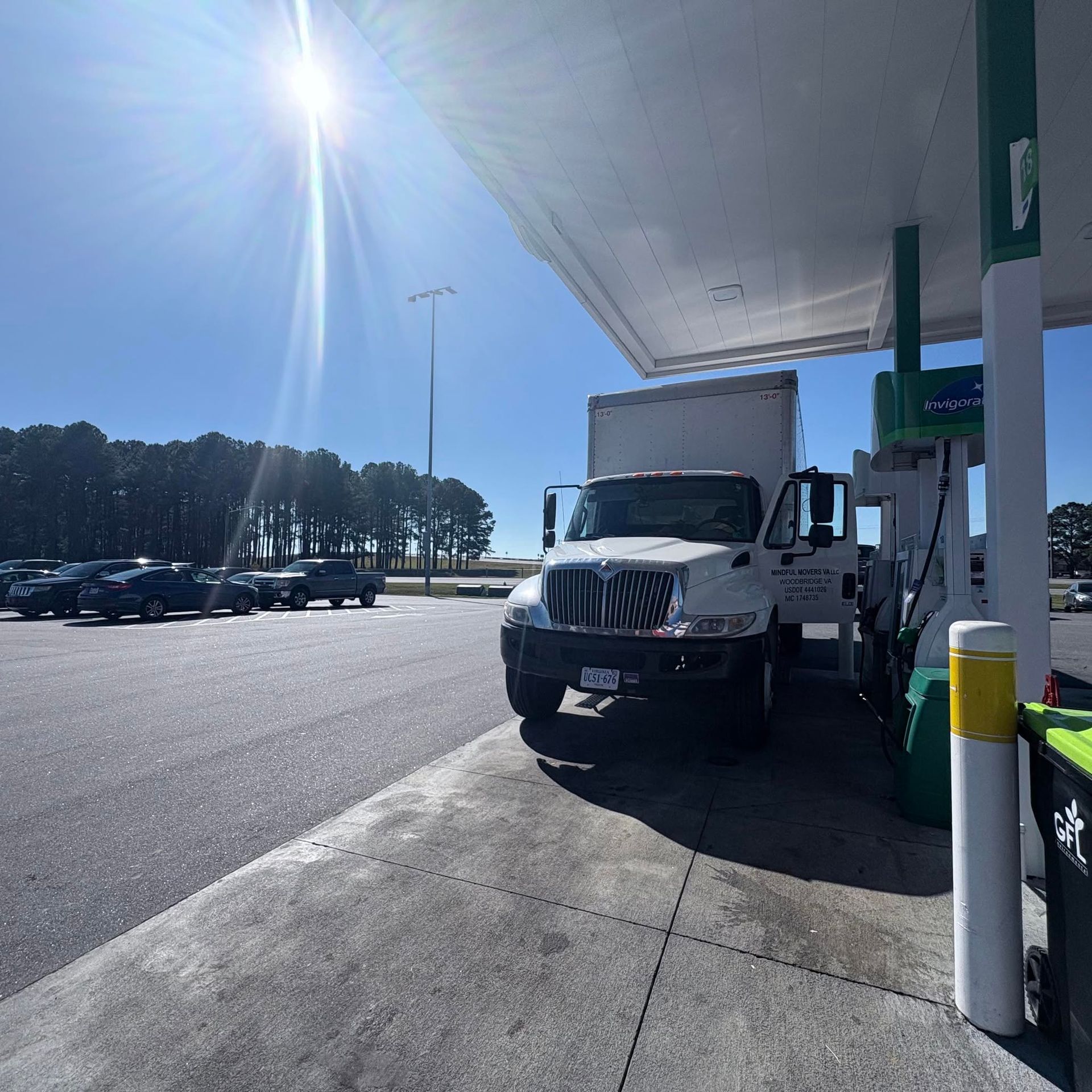 Semi truck parked at a gas station under a bright sun, with cars in the lot nearby.