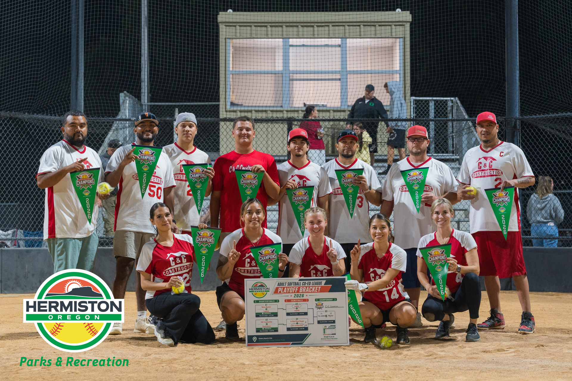Hermiston Parks & Recreation softball team posing on a field at night with a scoreboard behind them.