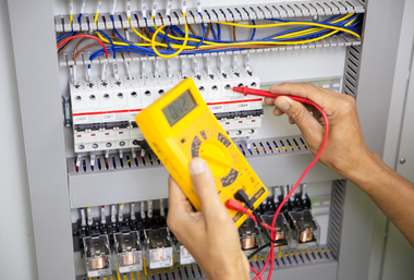 Hands testing electrical panel with yellow multimeter and red probes