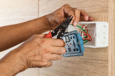 Hands wiring an electrical outlet in a wall box with pliers and exposed colored wires