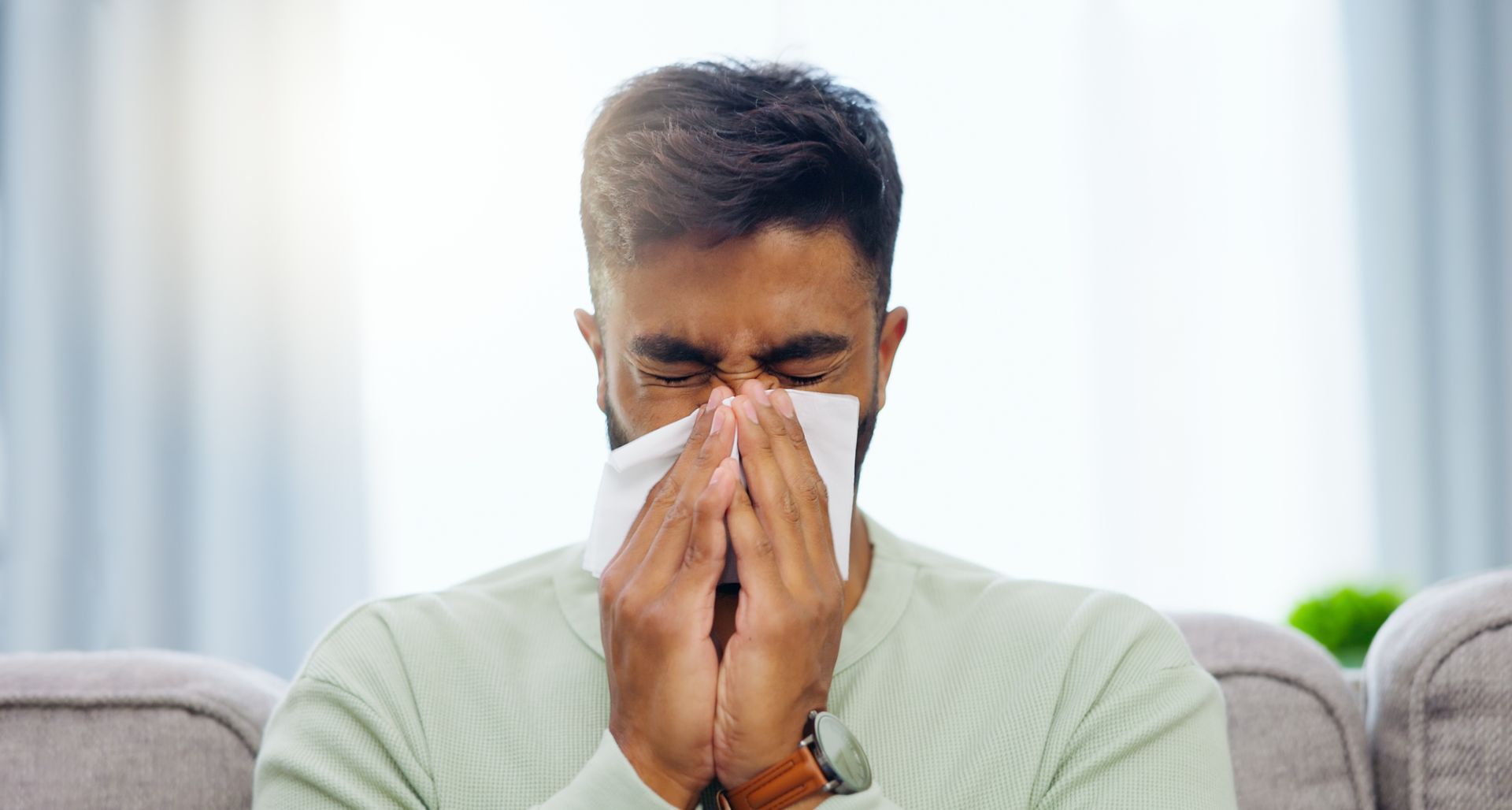 A man is blowing his nose into a napkin while sitting on a couch.