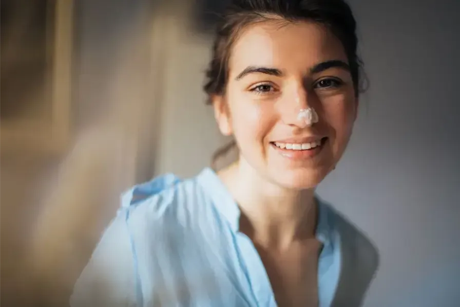 A smiling young woman with a dab of white moisturizing cream on the tip of her nose