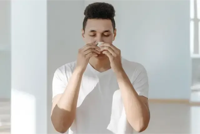 Person in a white shirt holding a tissue to their nose in a bright room