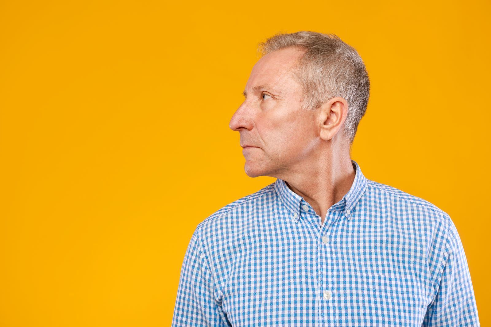Older man in a blue checkered shirt, looking left with a serious expression against a yellow background.