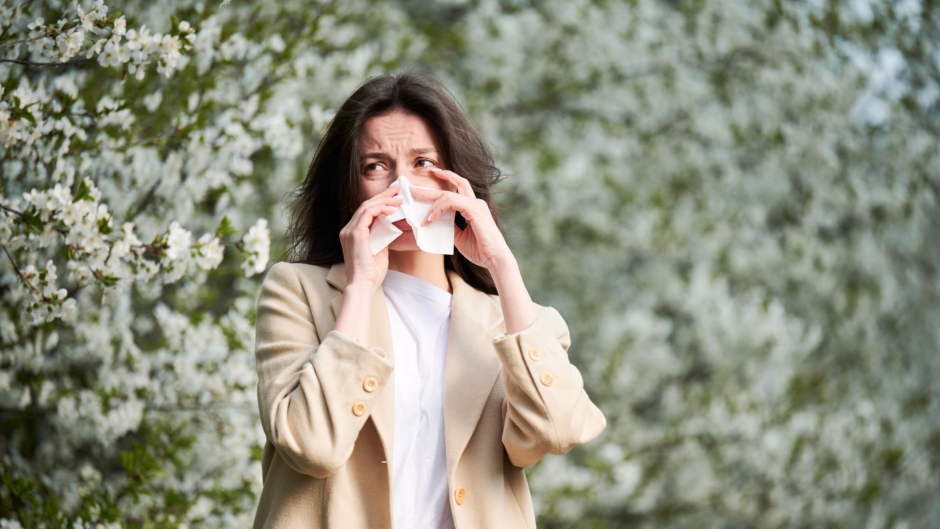 Woman in a tan blazer blowing her nose with a tissue, standing in front of a flowering tree. She has a pained expression.