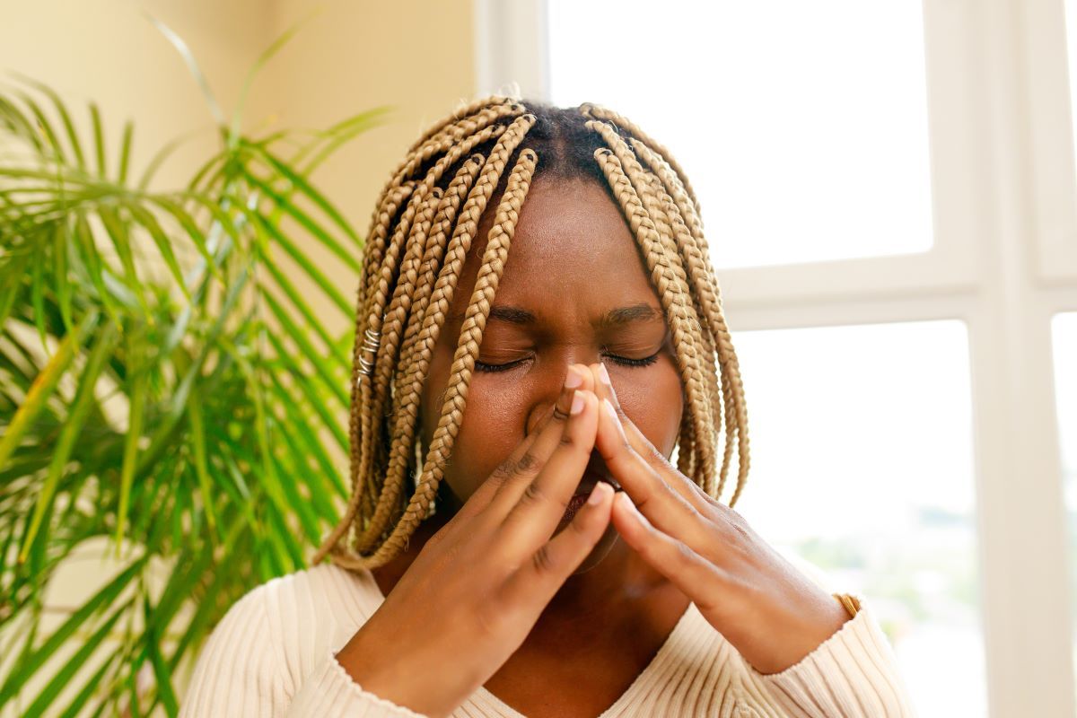 A young Black woman with blonde braids, eyes closed, presses her fingers against her nose. In a bright room with a plant.