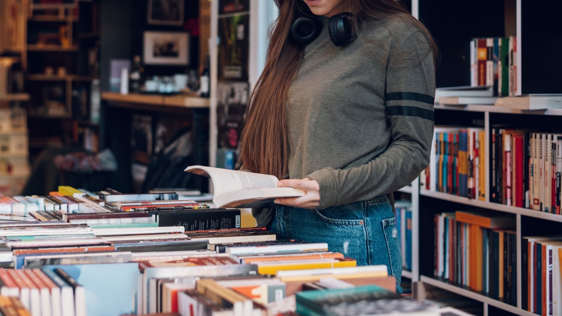 Person browsing books in a bookstore aisle, holding an open book near shelves and stacked books.