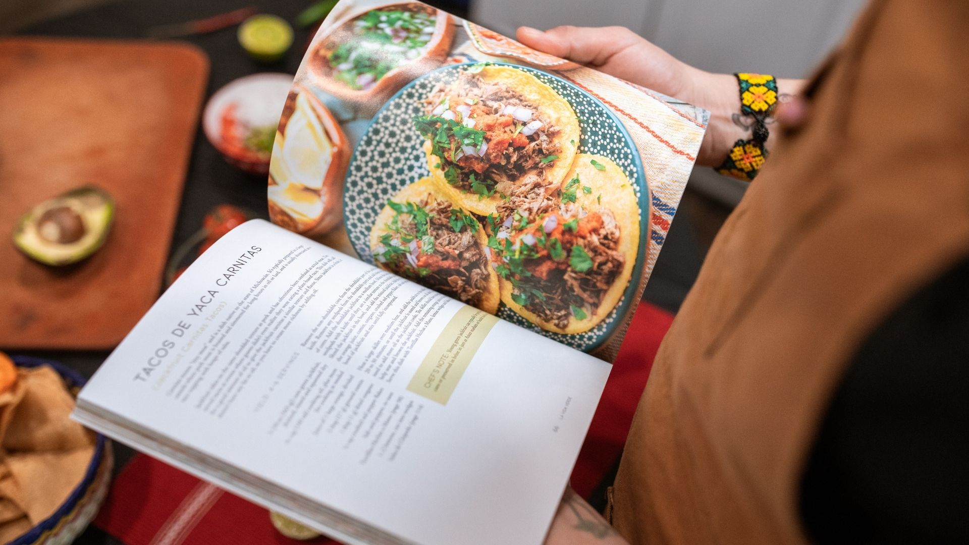 Person reading a colorful cookbook recipe with photos of plated dishes open on a table