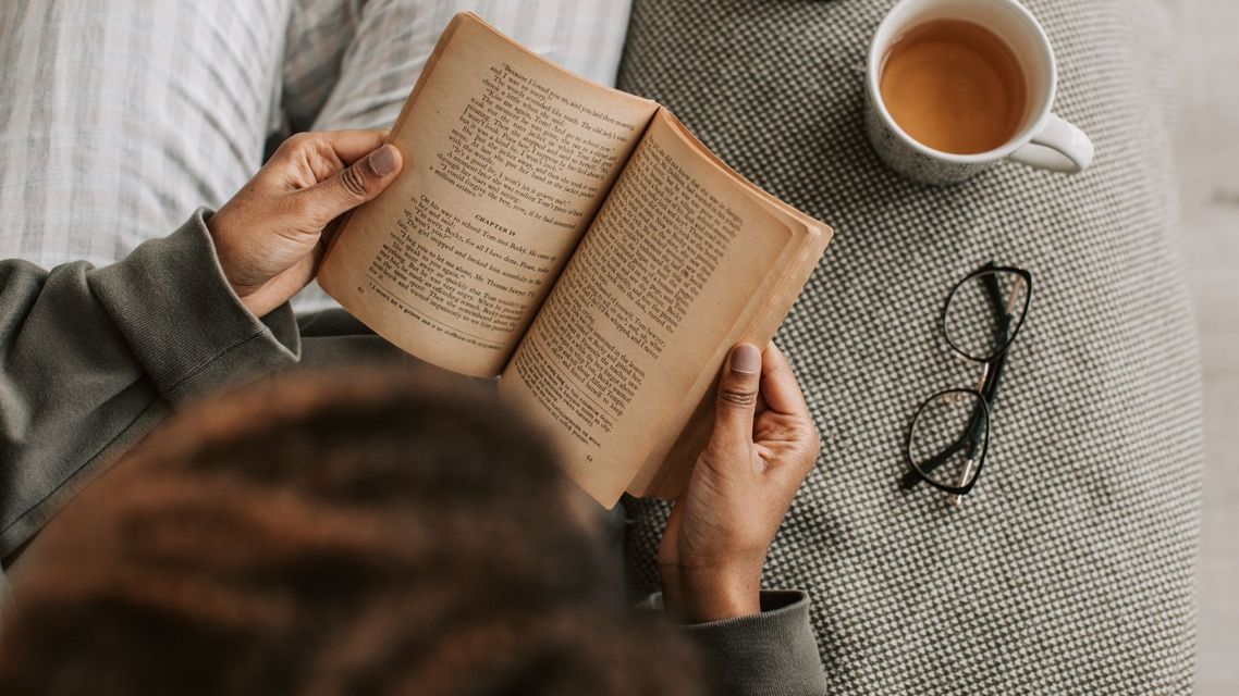 Person reading a book on a gray couch beside a cup of tea and glasses
