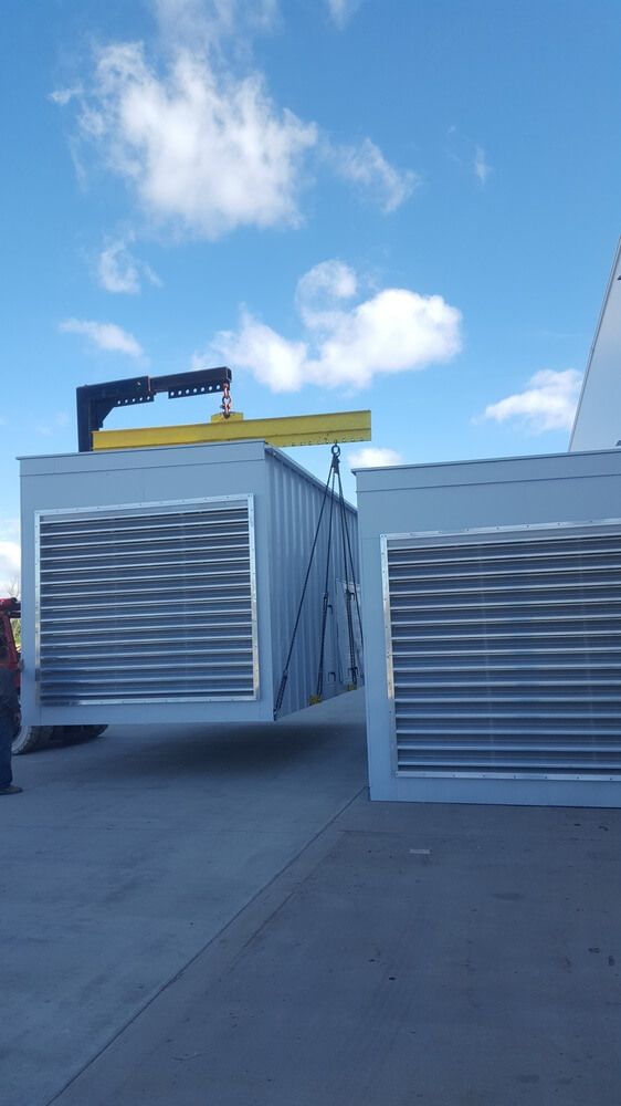 Crane lifting a large, rectangular industrial unit with slatted front, blue sky background.