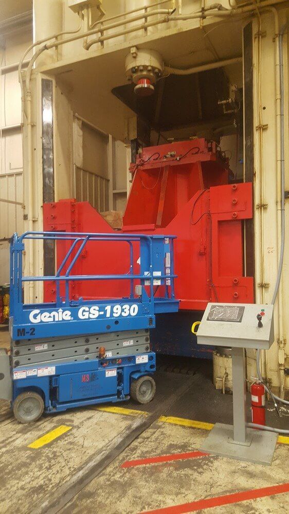 Large red industrial press with a blue lift in front; control panel and fire extinguisher are nearby.