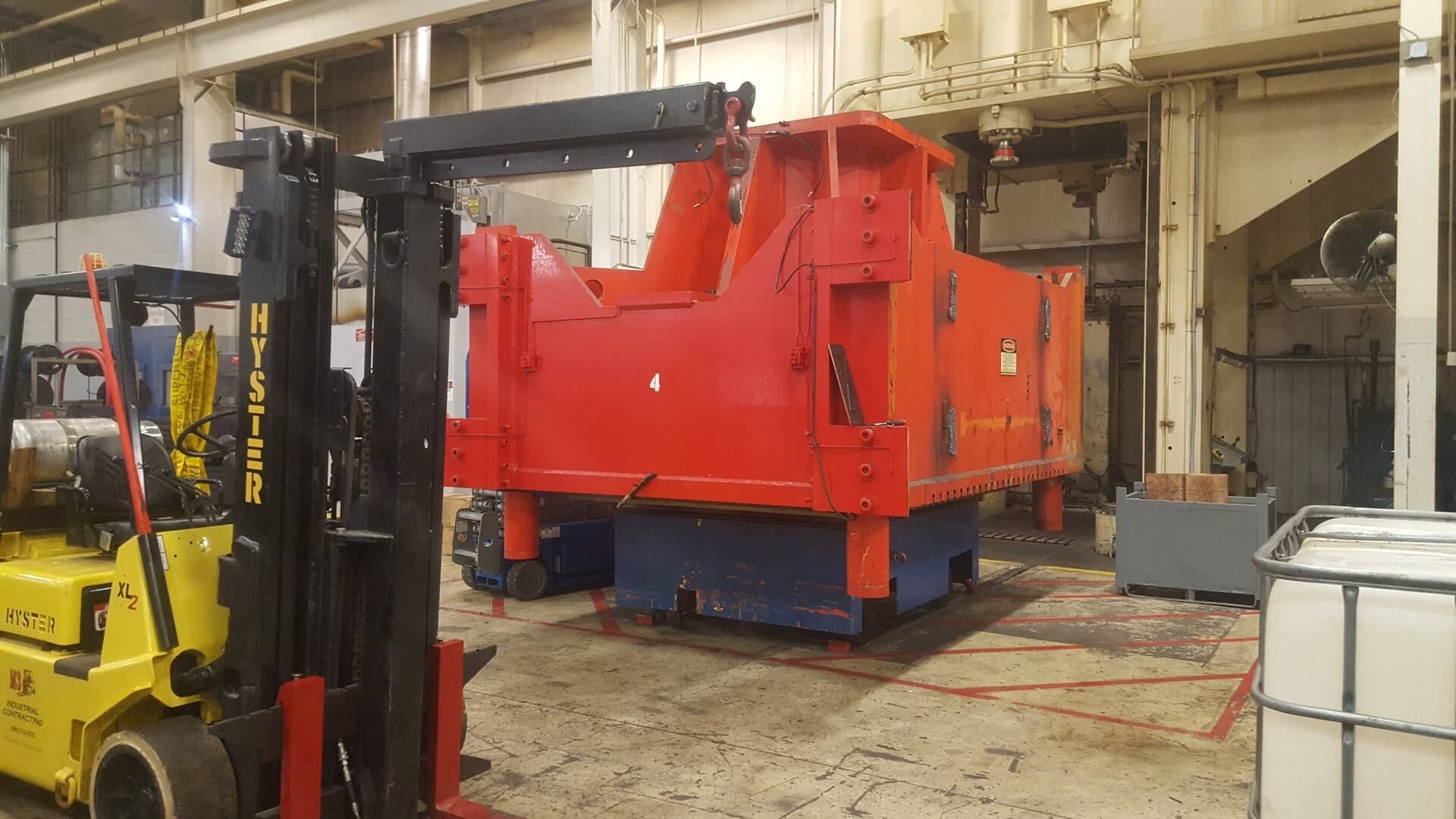 A yellow forklift lifting a large, red industrial machine in a factory setting.