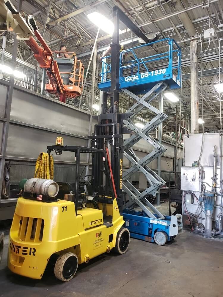 Yellow forklift and blue scissor lift in a factory setting, with a person in an aerial lift in the background.