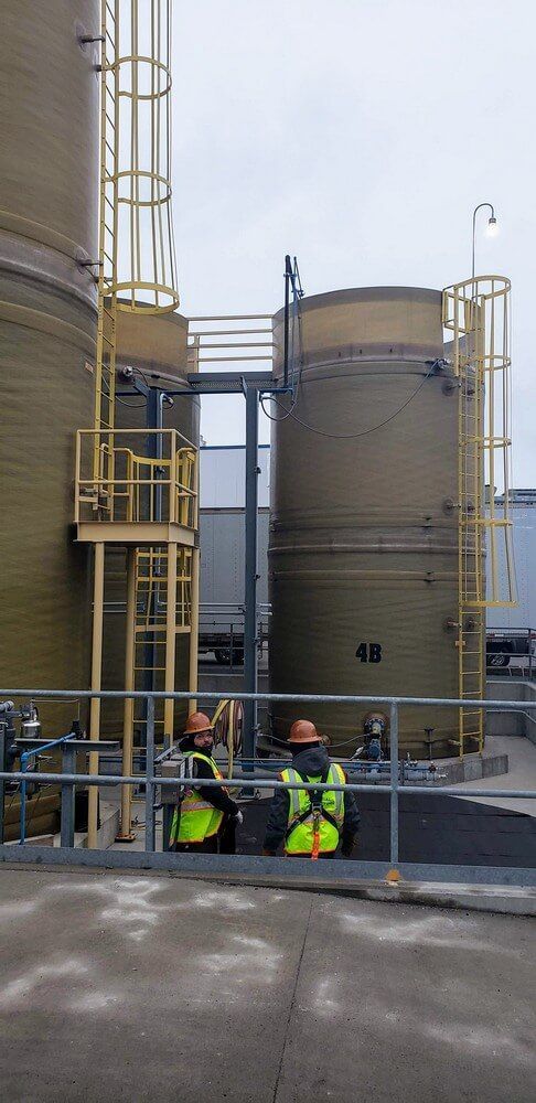 Two workers in safety gear near industrial tanks with ladders and railings.