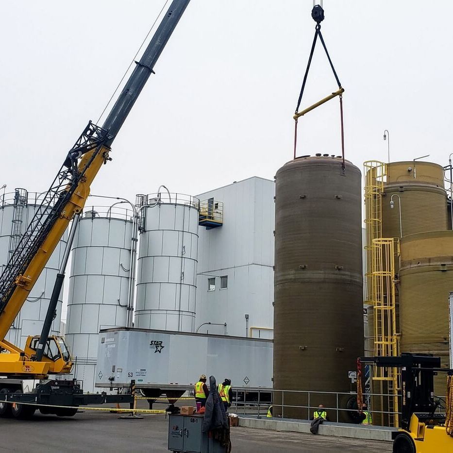 Crane lifting a large cylindrical tank at an industrial site; workers in yellow vests nearby.