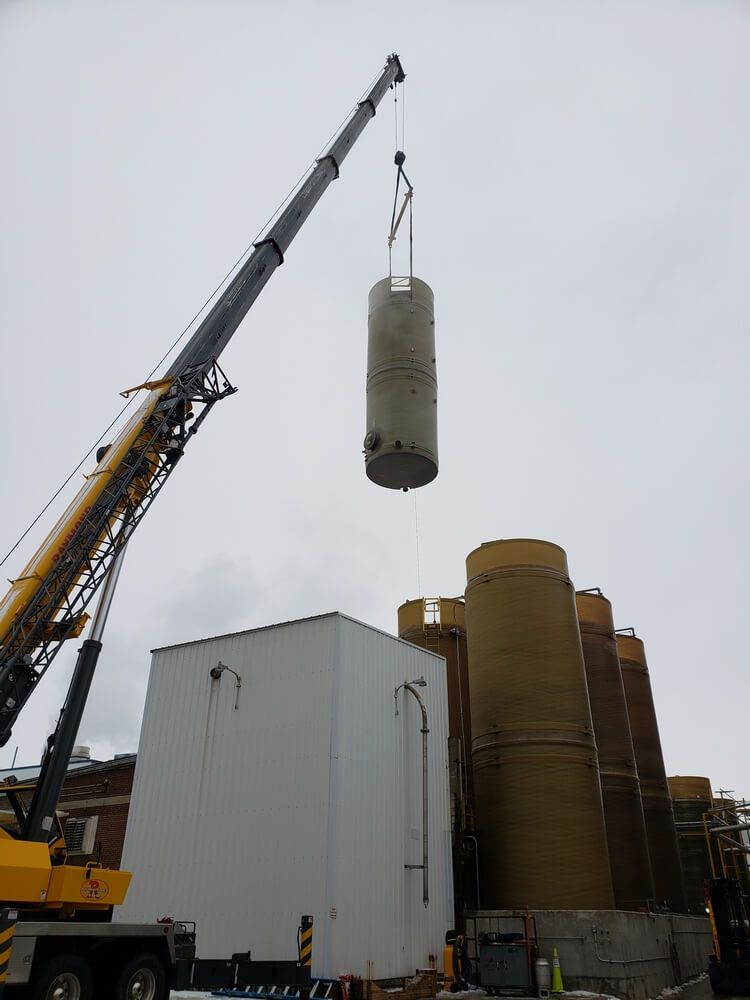 Crane lifting a cylindrical object. Beige object, white building, and yellow silos against cloudy sky.