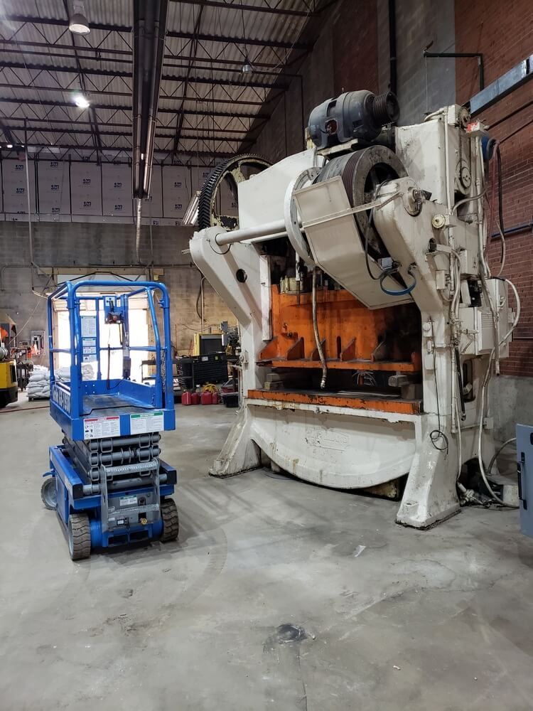 Blue scissor lift next to a large, white industrial press in a warehouse.
