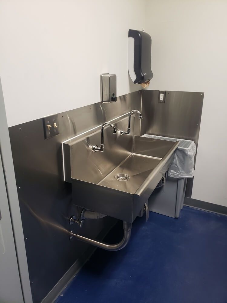 Stainless steel sink in corner with hand soap and paper towel dispensers. Blue floor, white walls.