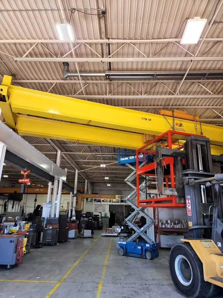 Yellow overhead crane in a large warehouse. A scissor lift and forklift are also present.