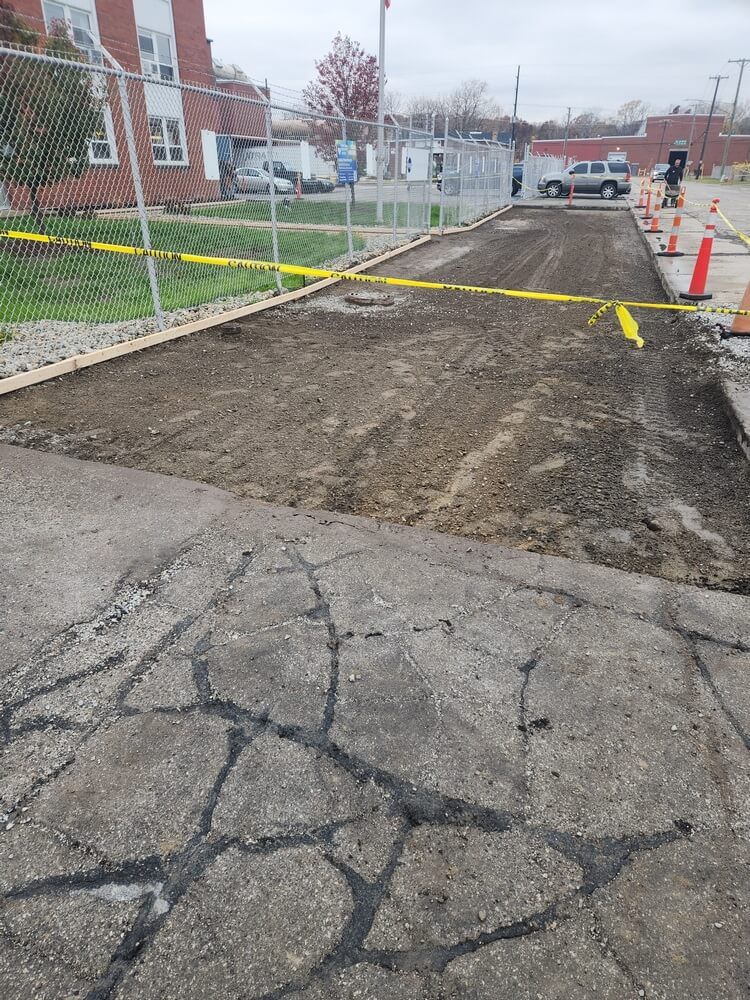 Paved area with section removed for repair. Yellow caution tape and construction cones. Faded building in the background.