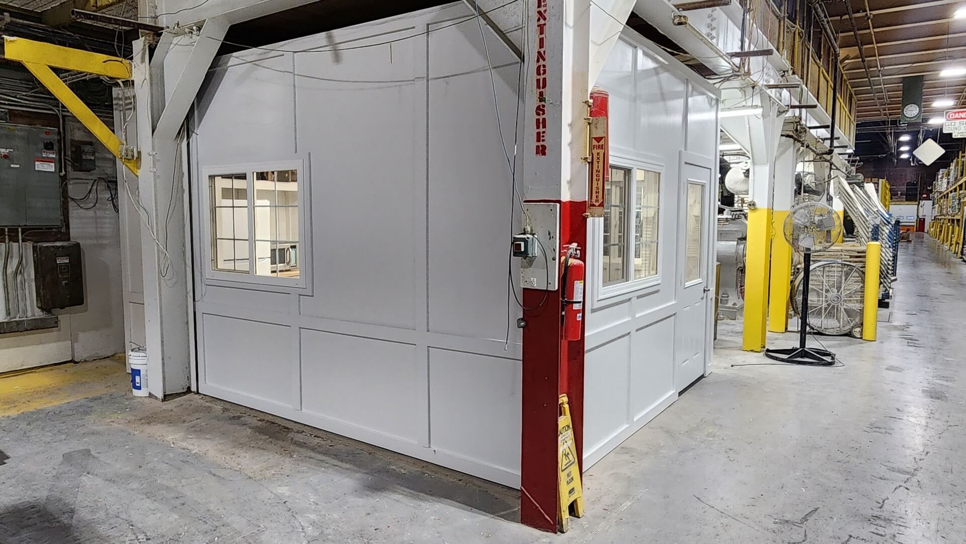 White industrial booth with windows in a factory setting. Red support beams, yellow safety lines, concrete floor.