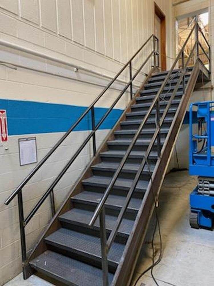 Metal staircase with black handrails and gray treads, against a white and blue wall.