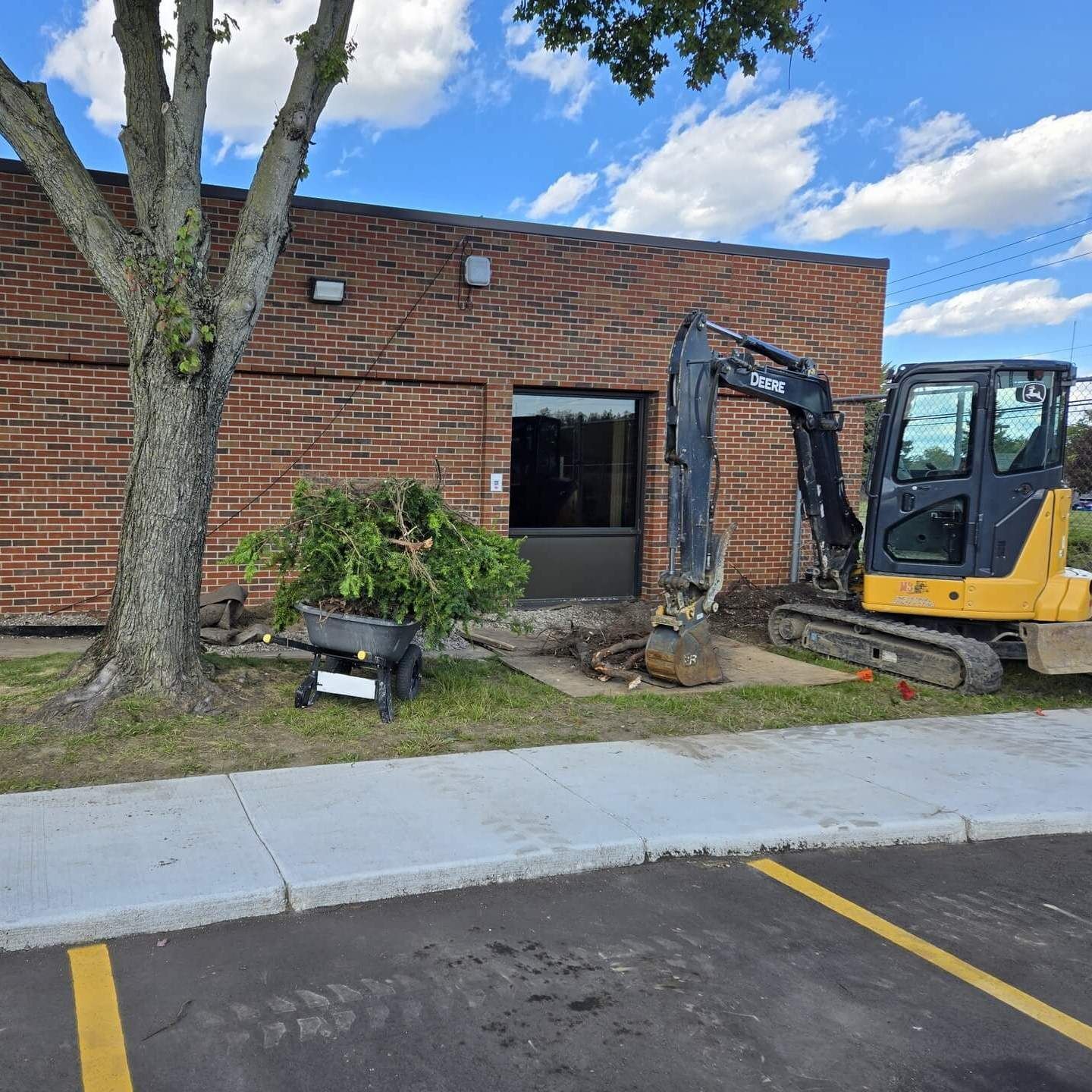 Construction site: excavator next to a brick building. Tree and debris on the lawn; blue sky.