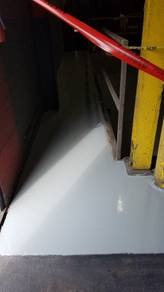 Light gray painted floor in an industrial setting, viewed from an entryway. Red barrier and yellow support beams.