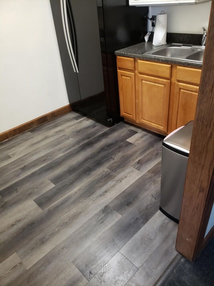 Kitchen with grey wood-look flooring, light wood cabinets, black refrigerator, stainless steel trash can and sink.