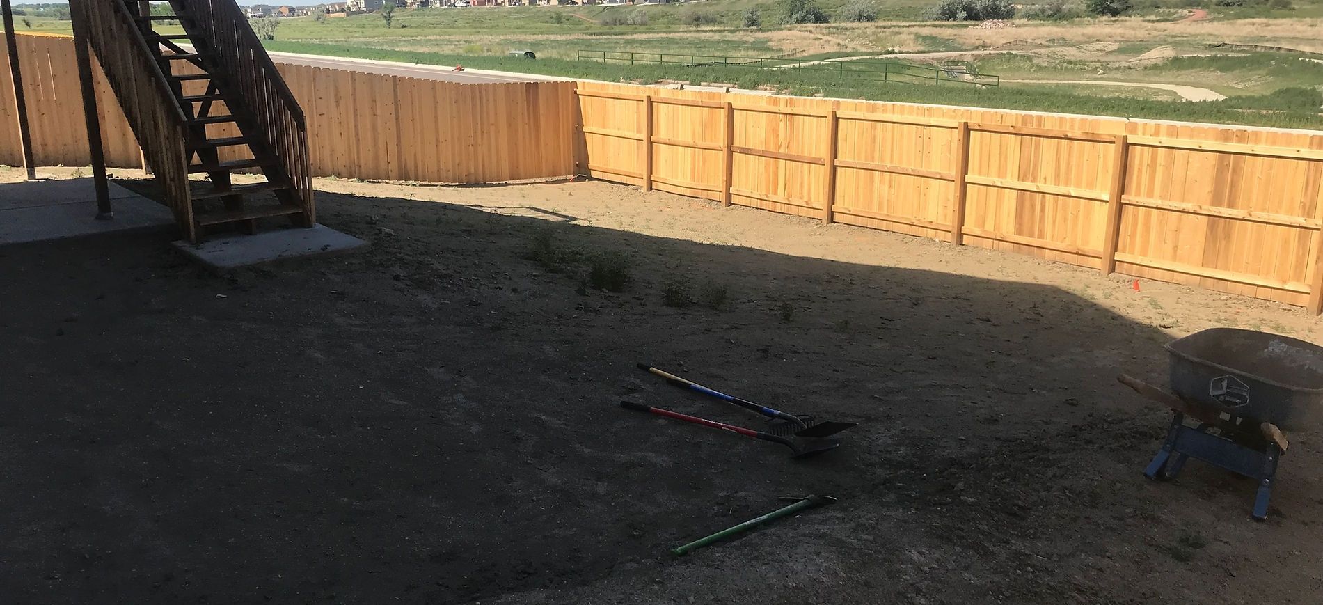 Backyard with a wooden fence, dark patio, and stairs casting shadows in sunlight