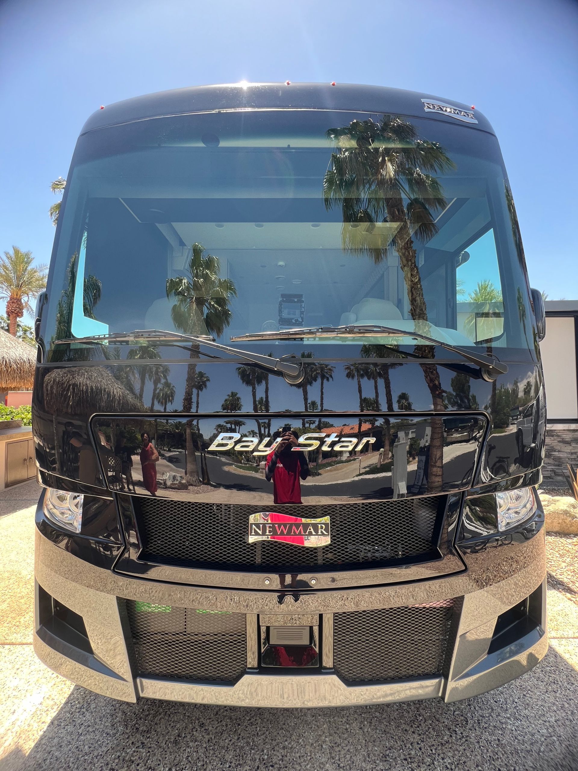 A black rv is parked in a gravel lot with palm trees reflected in the windshield.