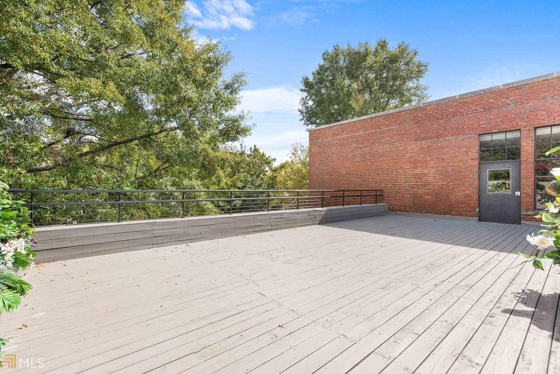 Empty rooftop deck with gray boards, black railing, and red brick wall beside trees and blue sky