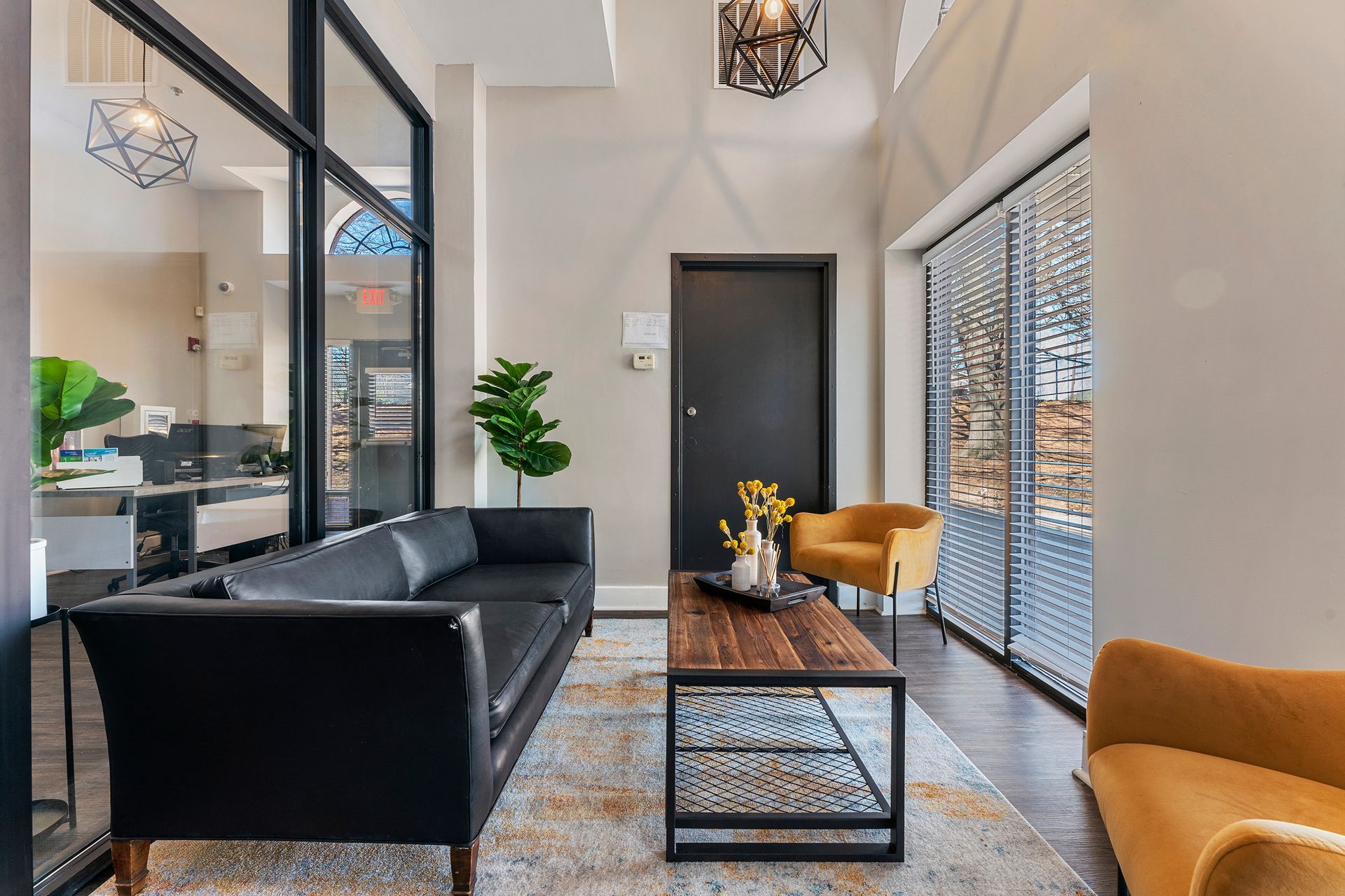 Modern living room with black sofa, mustard chairs, glass coffee table, and tall windows with blinds.