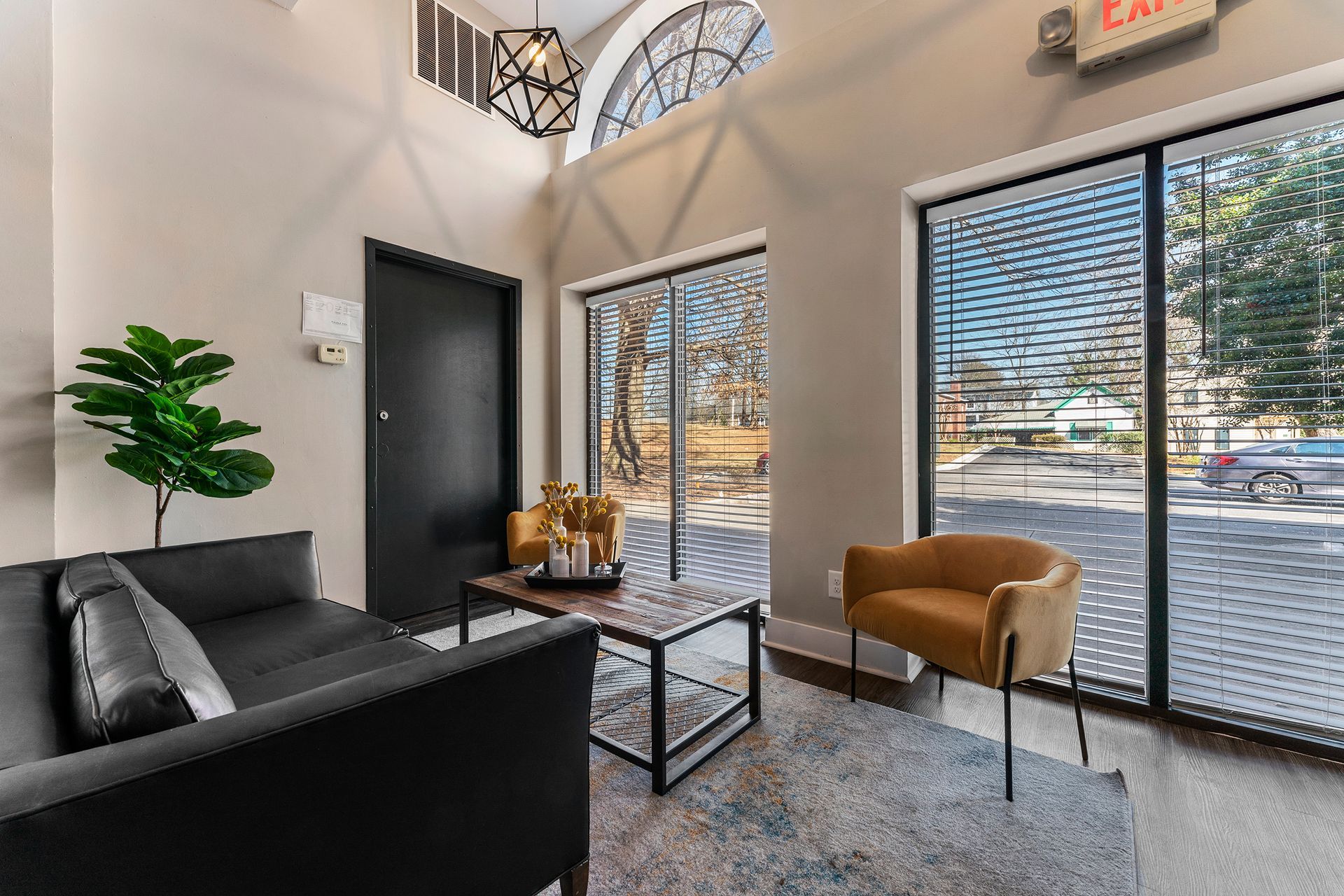 Modern waiting area with black sofa, tan chair, glass table, and large windows with blinds