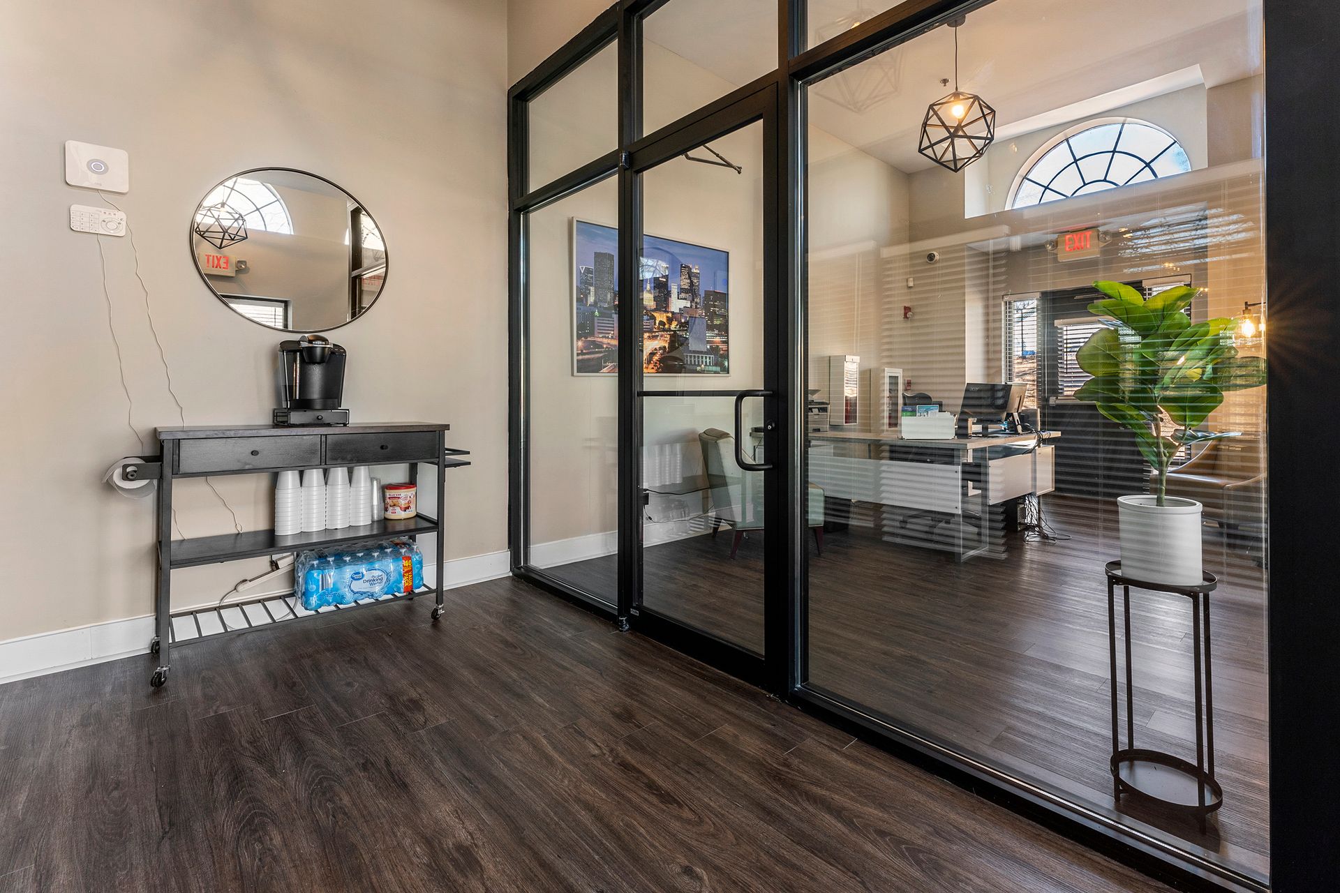 Modern office lobby with glass doors, dark wood floors, a console table, mirror, and potted plants.