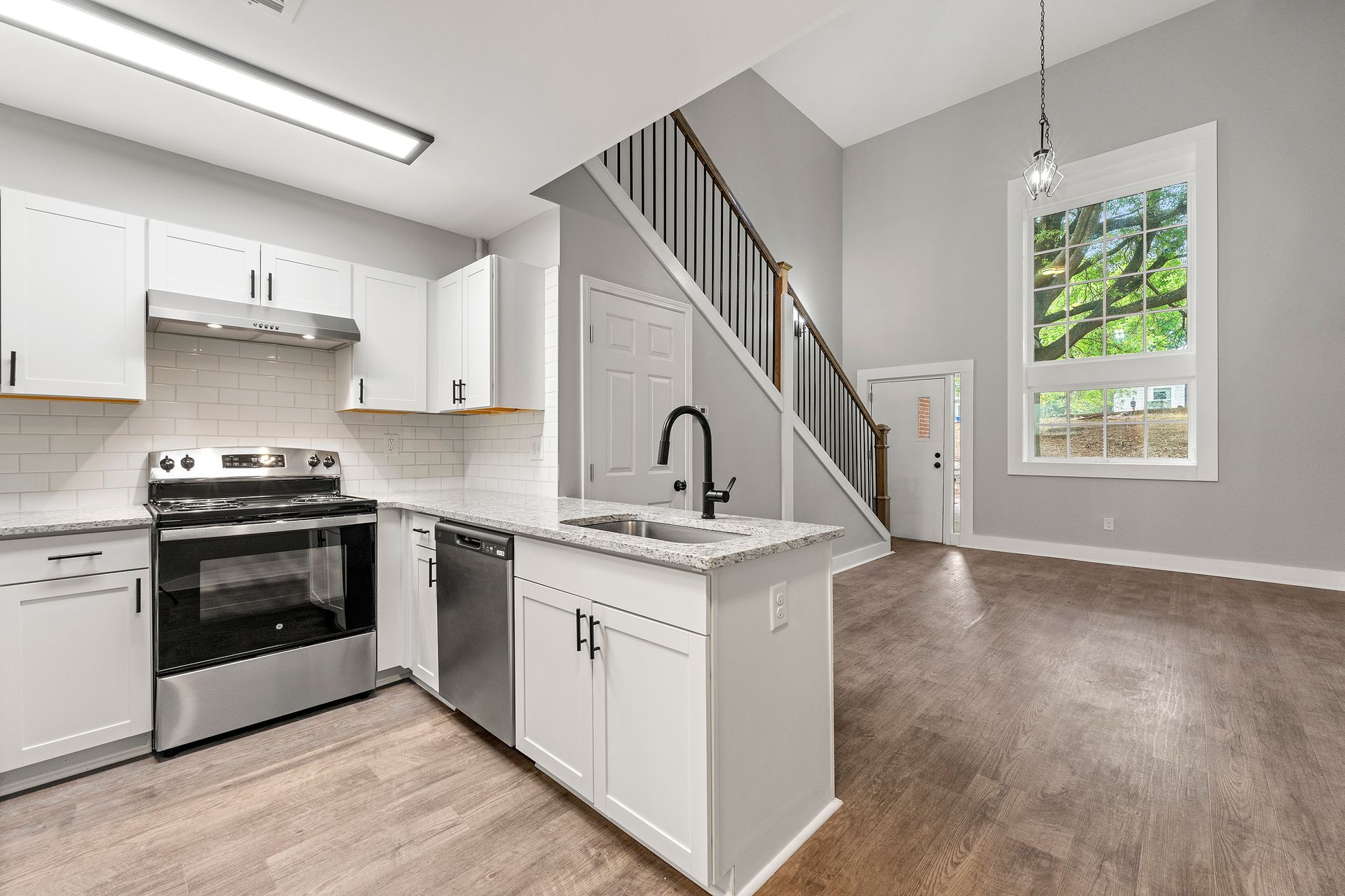 Bright kitchen with white cabinets, stainless stove, island, and staircase beside a carpeted living area.