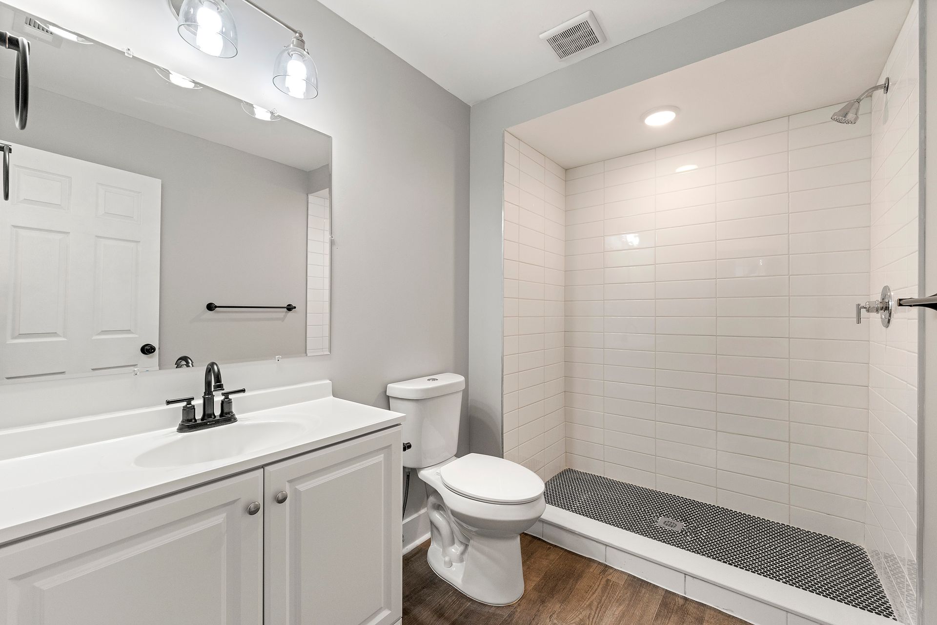 Bright bathroom with white vanity, toilet, mirror, and tiled walk-in shower with black mosaic floor.
