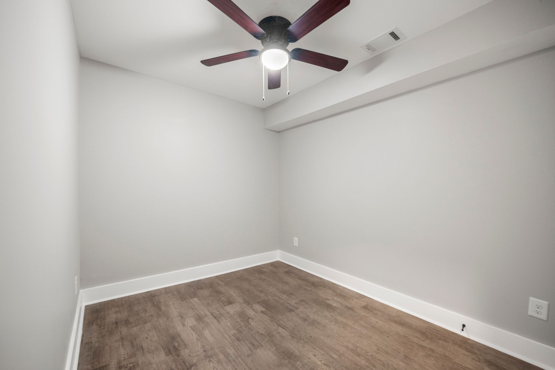Empty gray bedroom with white trim, wood floor, and a ceiling fan with light fixture.