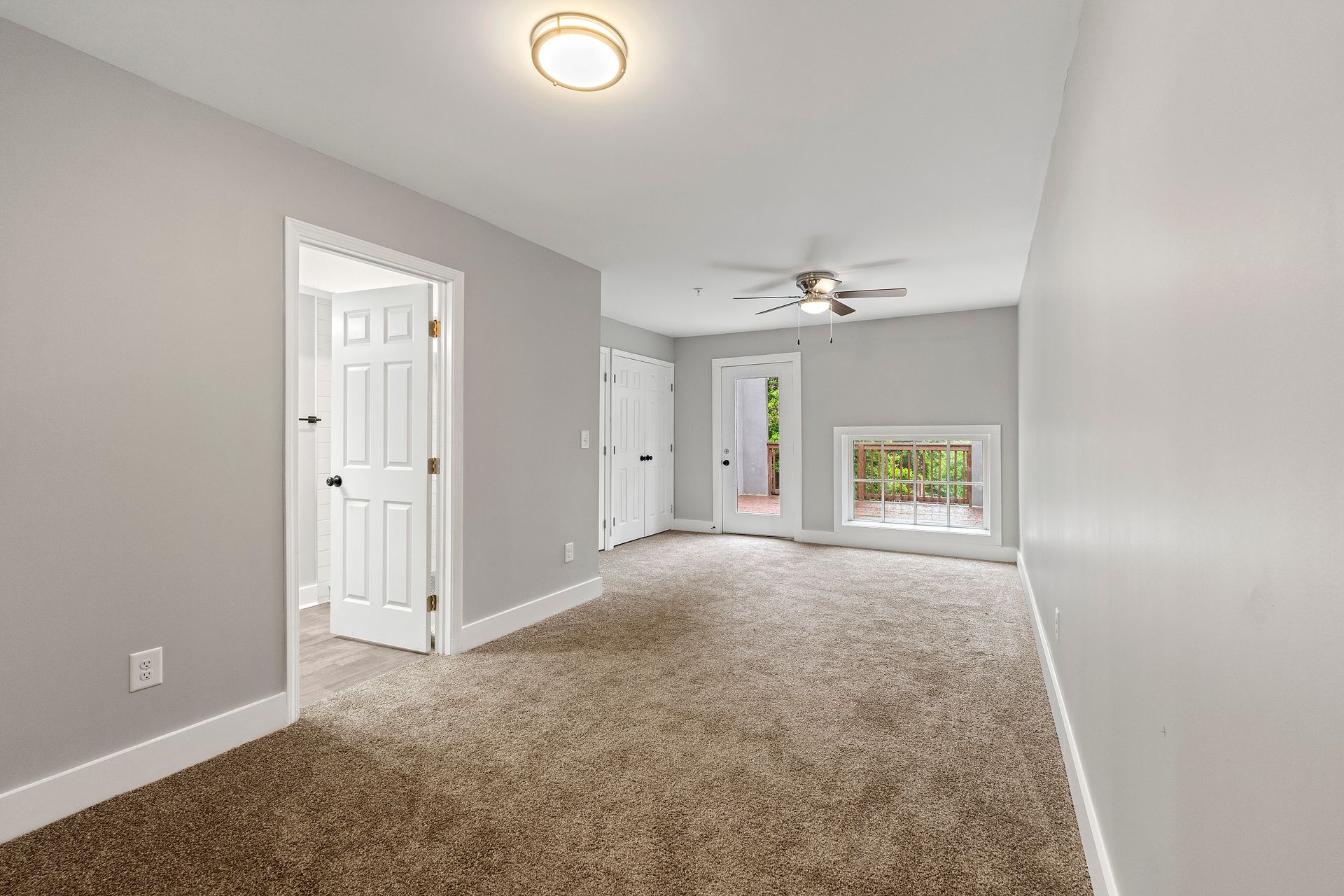 Empty carpeted living room with gray walls, ceiling fan, white doors, and a window.