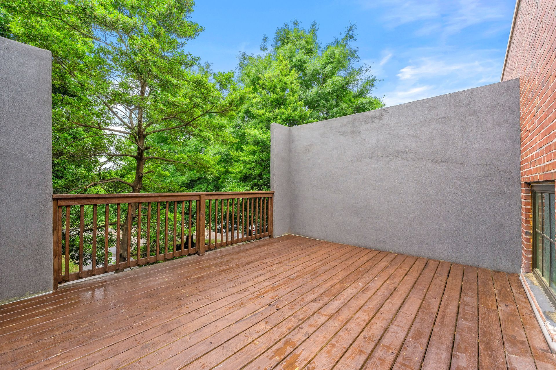Empty wooden deck with railing, gray privacy walls, and green trees in the background.