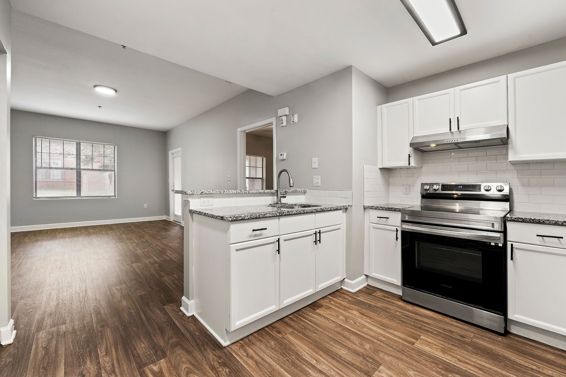 Bright modern kitchen with white cabinets, stainless steel stove, and wood-look floors, open to a living area.