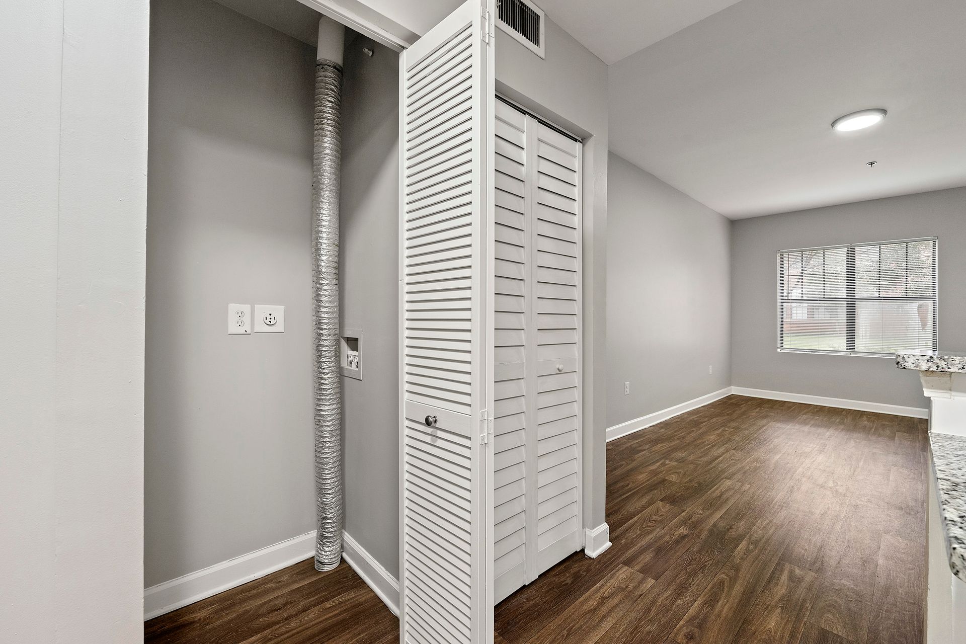 Empty gray apartment hallway with wood floors, louvered closet doors, and a window at the end.