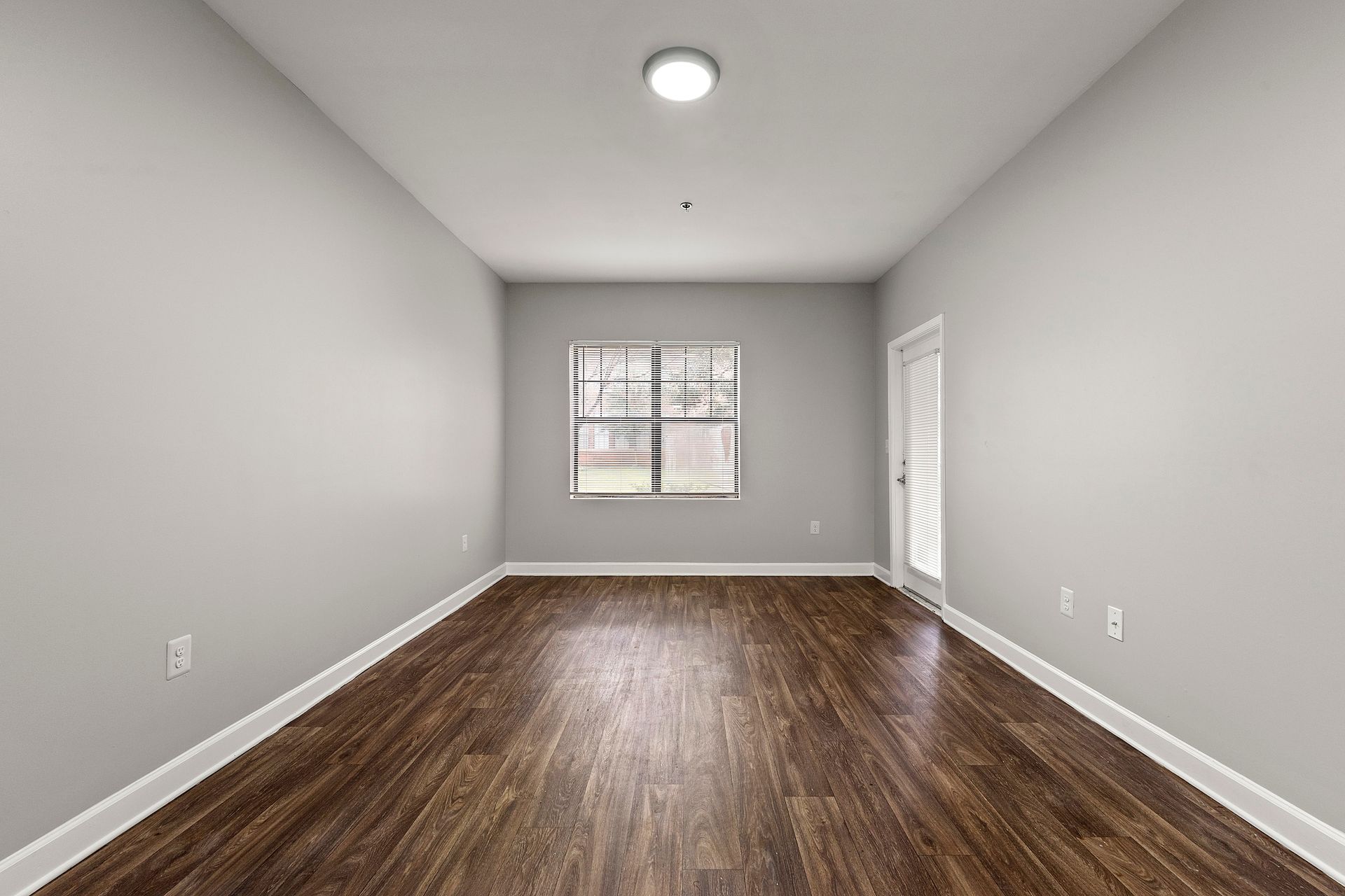 Empty gray room with dark wood floor, white ceiling light, and a window at the far end.