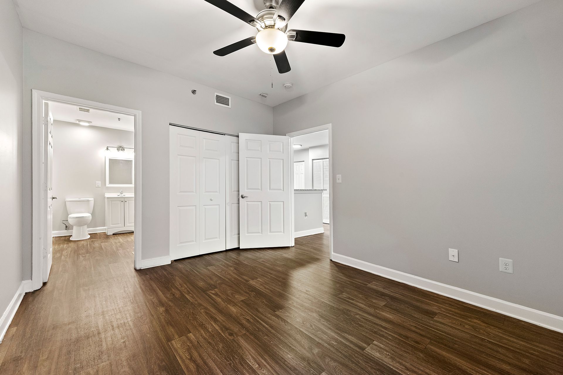 Empty bedroom with gray walls, wood floors, ceiling fan, and open door to a bathroom.