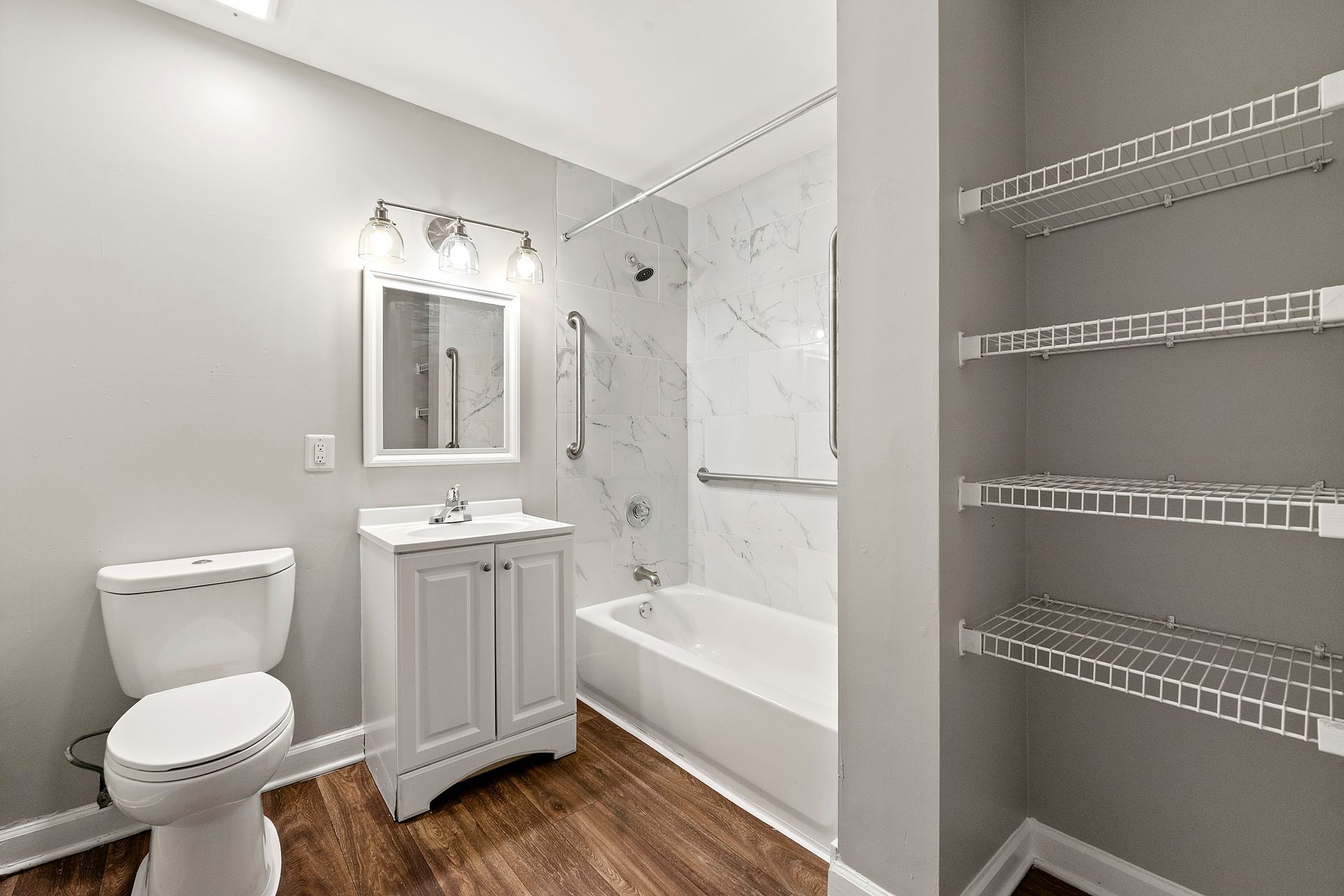 Small bathroom with white vanity, toilet, tub, and open wire shelving on the right.