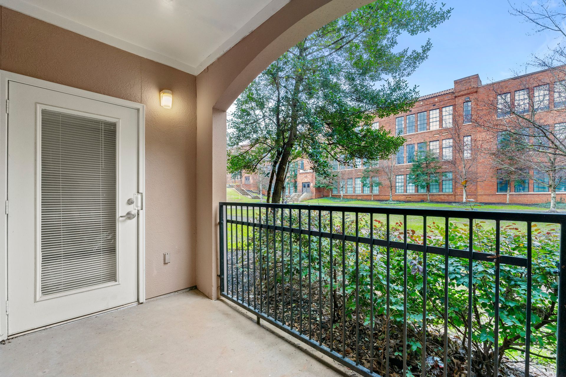 Apartment balcony overlooking trees and brick buildings, with black railing and white door.
