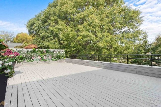 Spacious gray deck with black railing, potted flowers, and a large green tree under a blue sky