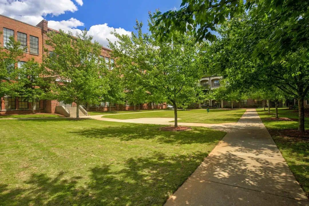 Tree-lined campus walkway beside a brick building and grassy courtyard on a sunny day