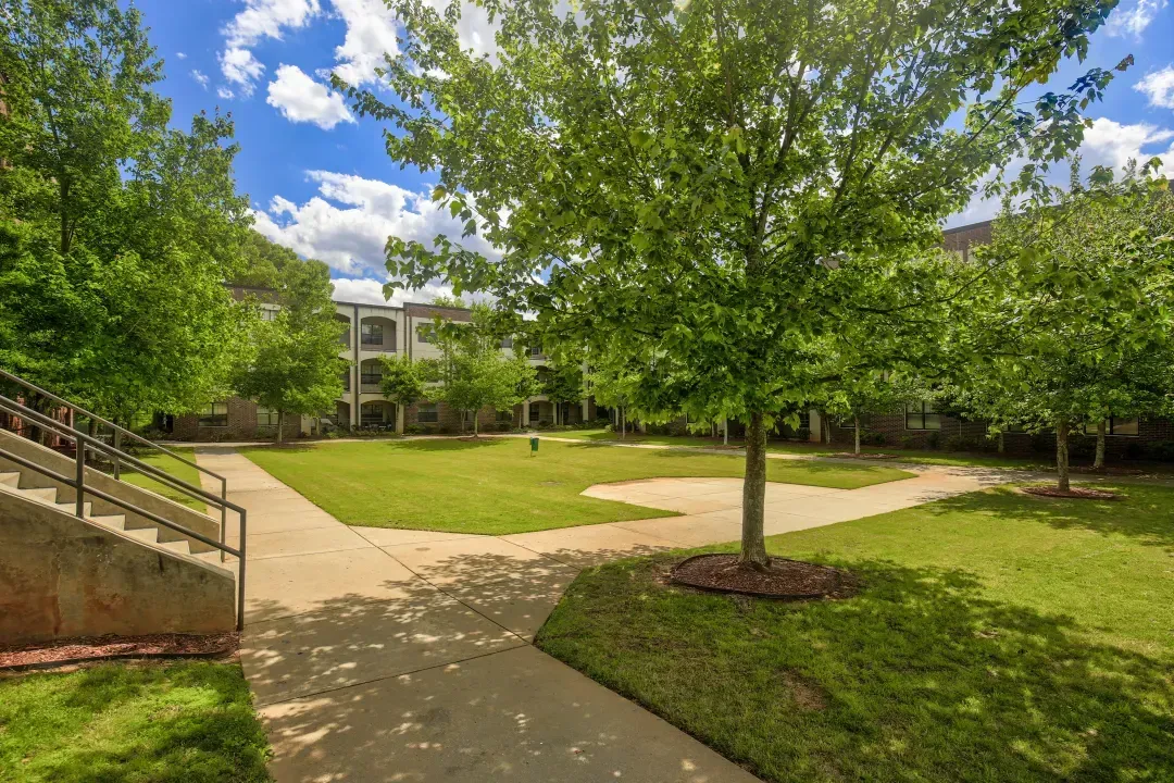 Tree-lined courtyard with grass, paved paths, and a building under a partly cloudy sky