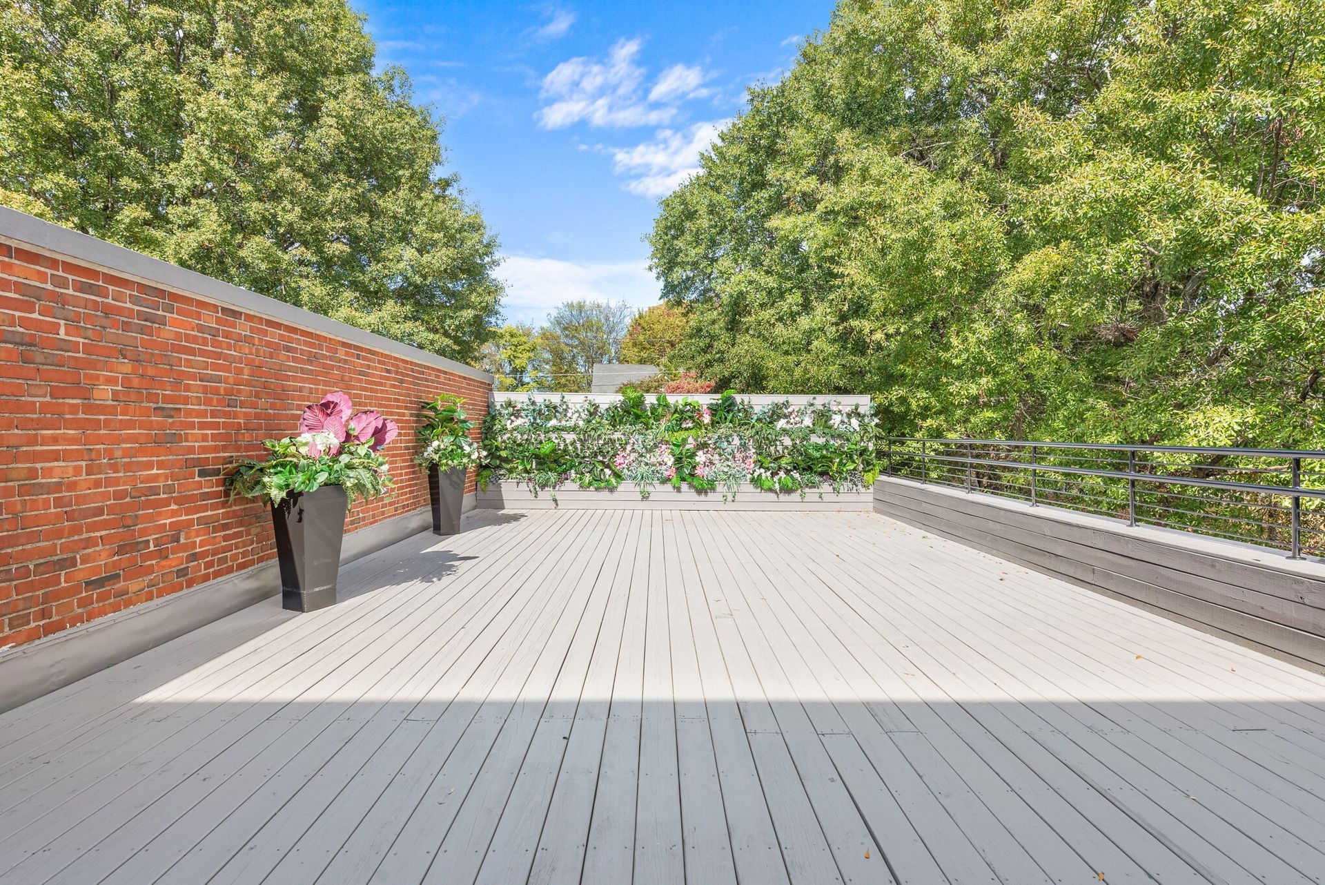 Spacious rooftop deck with light gray flooring, brick wall, potted flowers, and trees beyond the railing